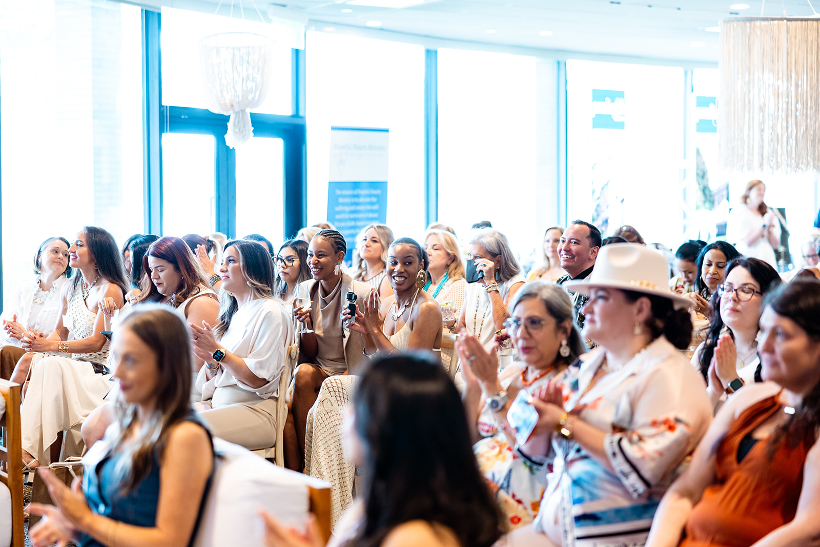 Group of people seated indoors near large windows, some clapping and smiling, bright natural light, modern setting.