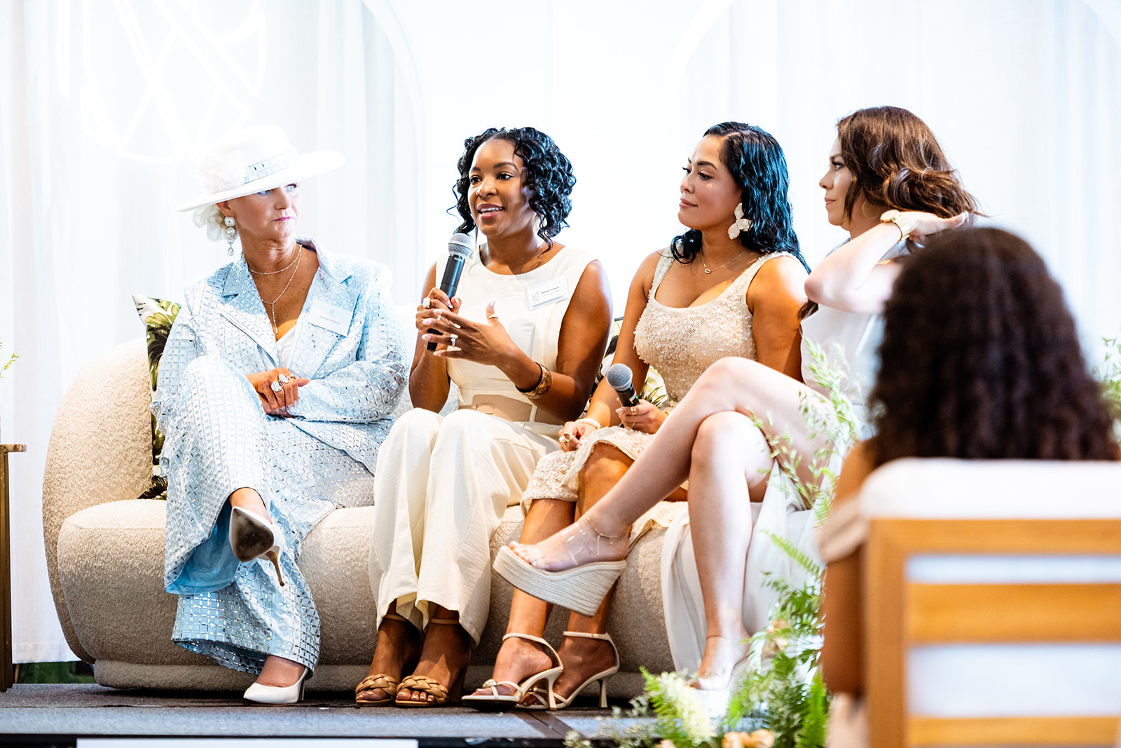 Five women sitting on a sofa, engaged in conversation, with a bright, airy background and plants nearby.