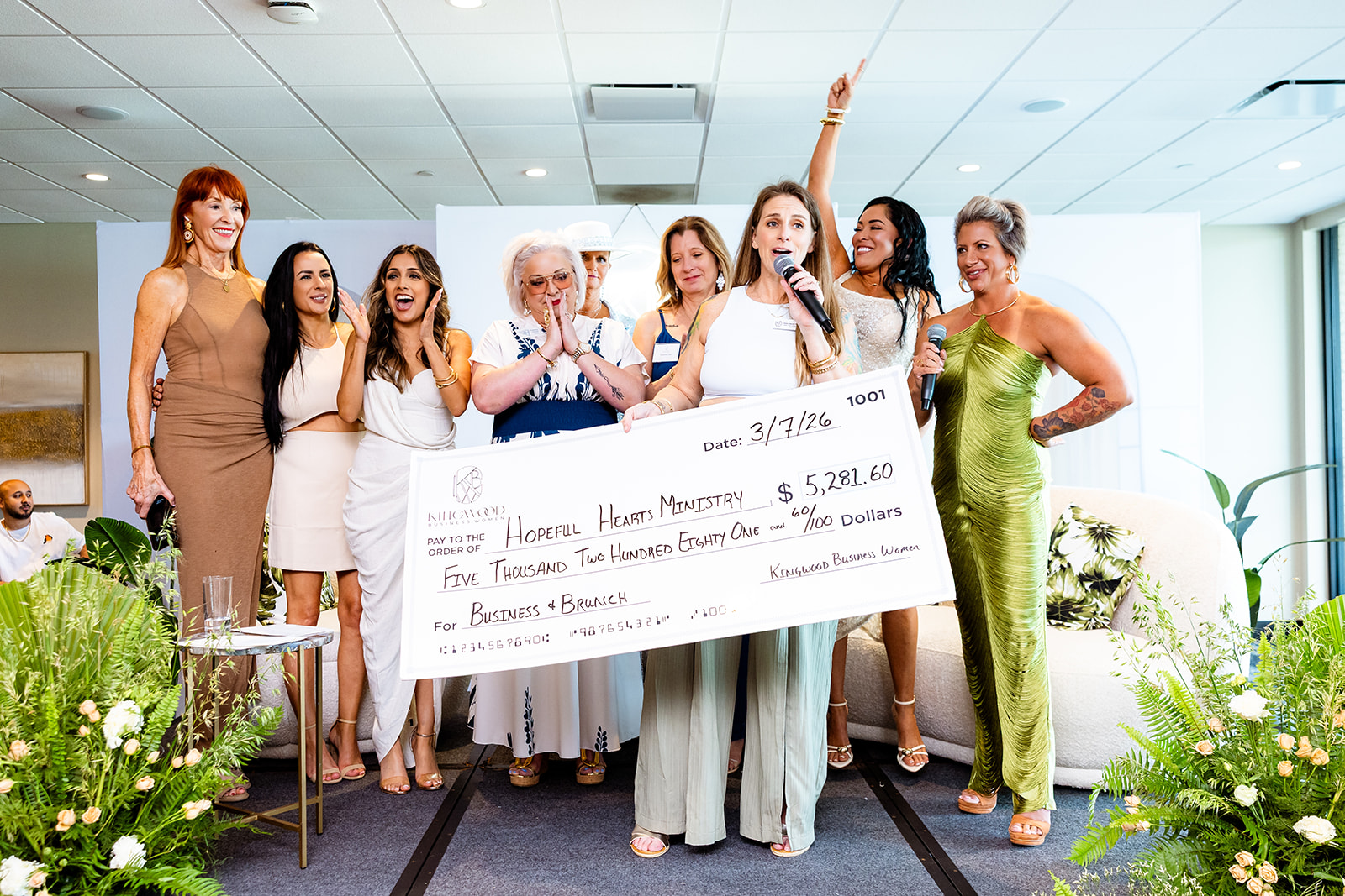 Group of women holding a large check in a bright room with plants and white furniture, celebrating.