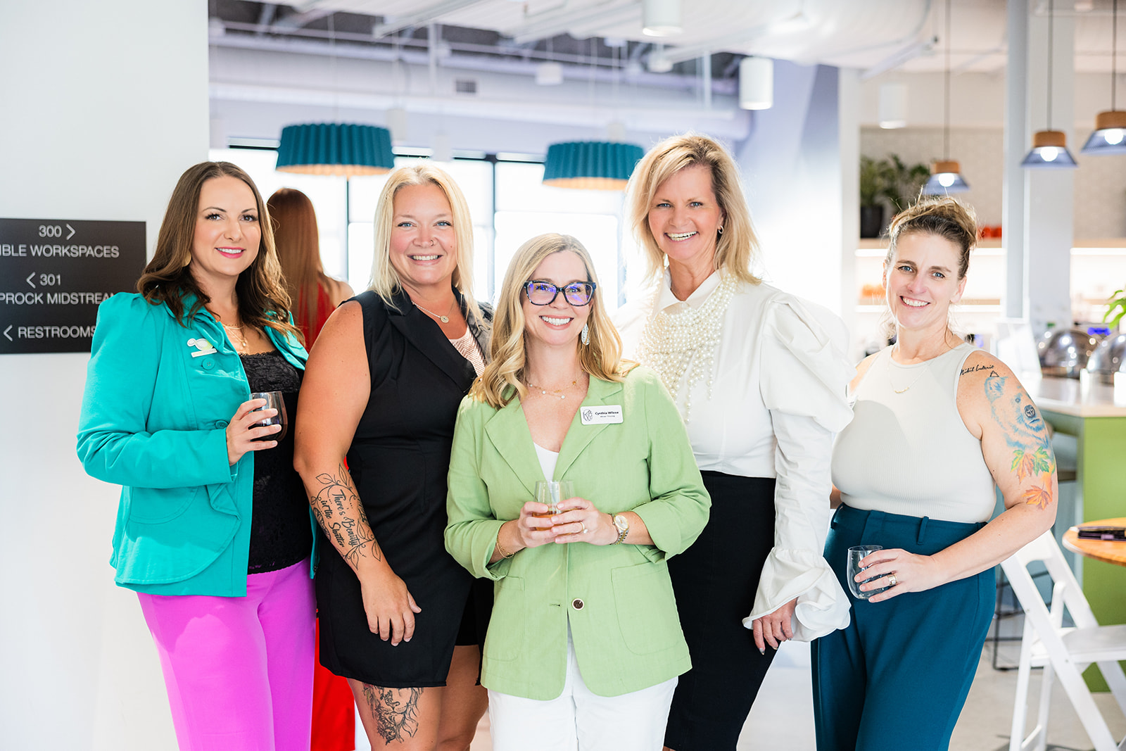 Group of five women smiling in a modern office space, some holding drinks, with bright lighting and contemporary decor.