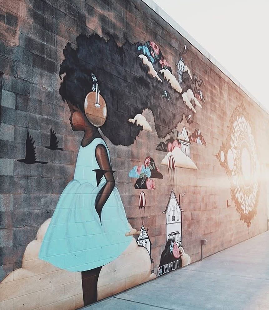 Mural of a girl with curly hair in a blue dress, clouds, and houses on a brick wall, viewed sideways.