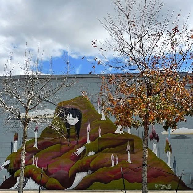 Street art mural of a girl with glasses and long hair on a hill, with trees and cloudy sky in background.