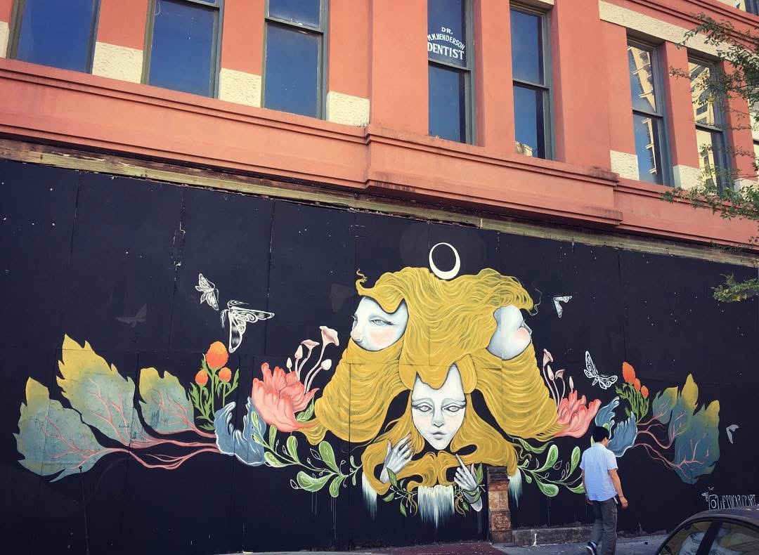 Street art mural of a woman with long yellow hair surrounded by colorful flowers, with a person standing nearby.