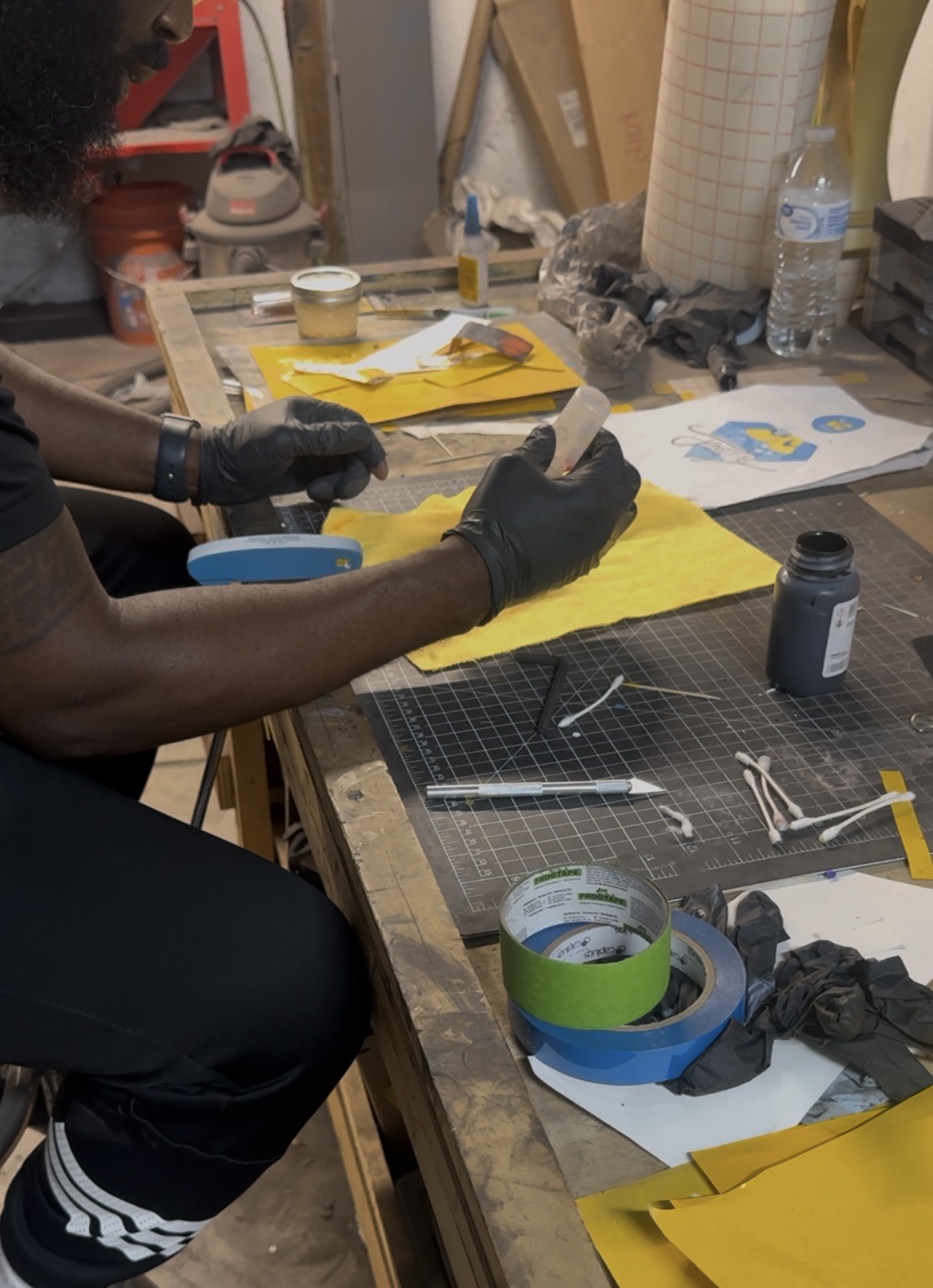Person working at a cluttered workbench with tools, tape, and paper, wearing black gloves and holding a small object.