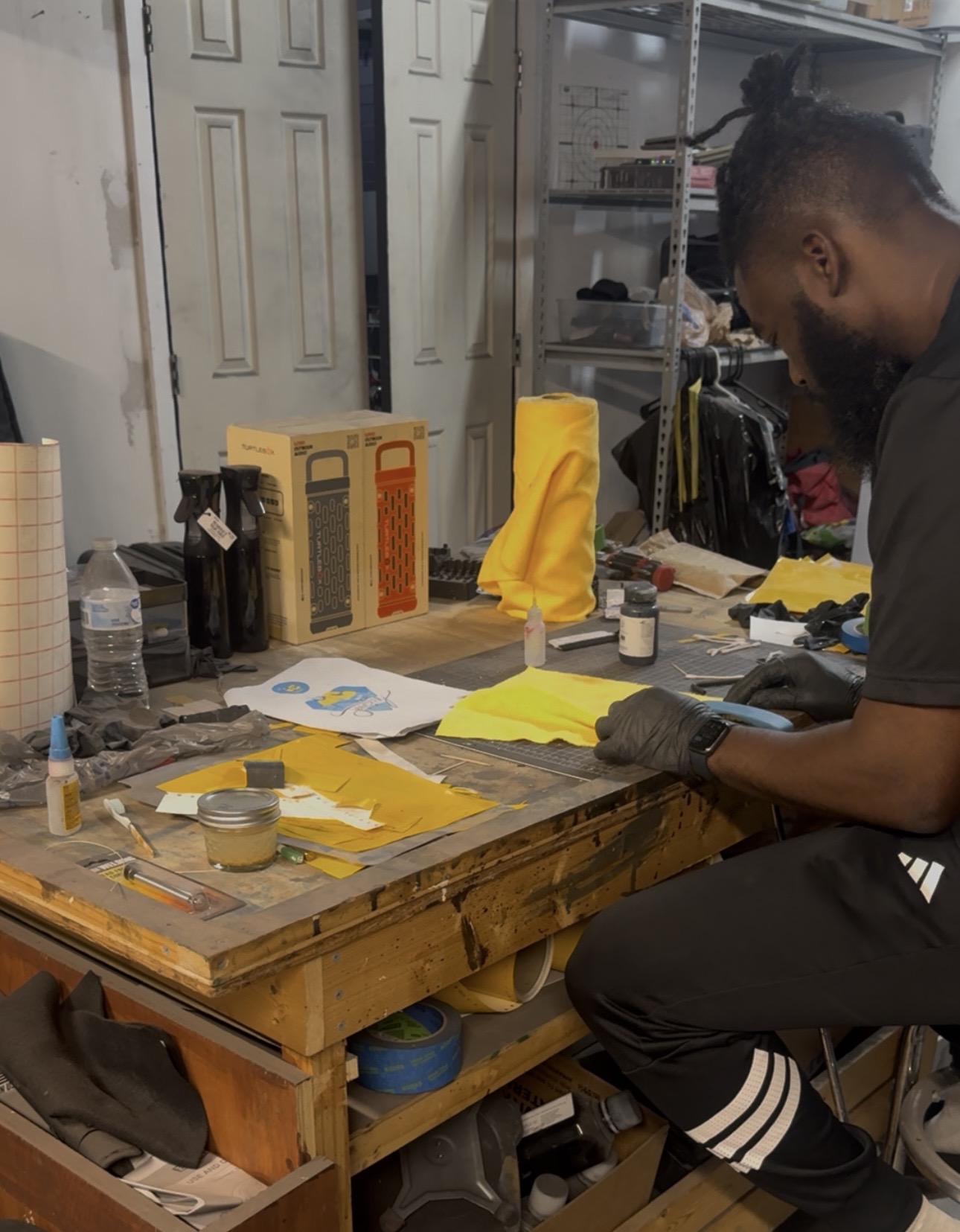 Person working at a cluttered workbench with yellow fabric, tools, and supplies in a workshop.