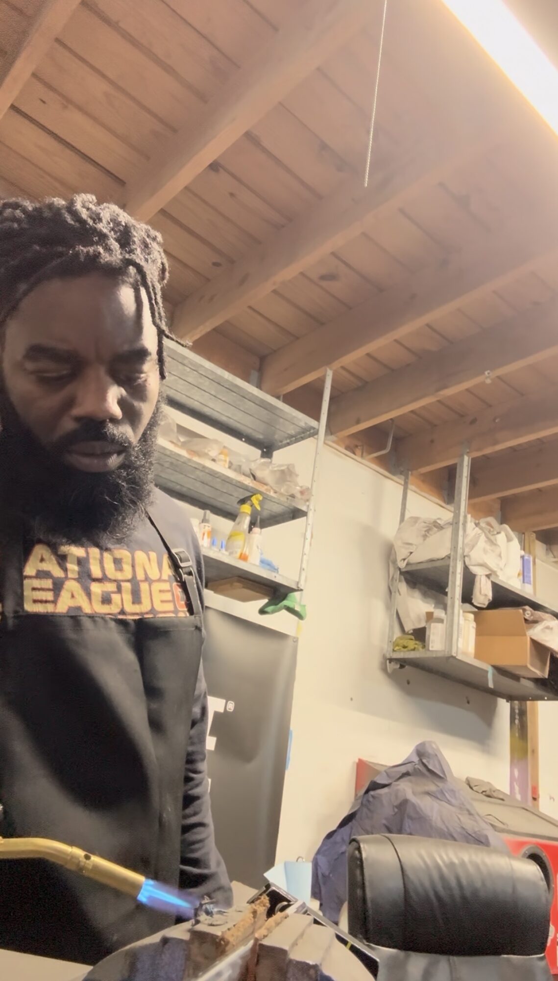 Man with dreadlocks wearing a black shirt with yellow text, standing in a cluttered room with a wooden ceiling.