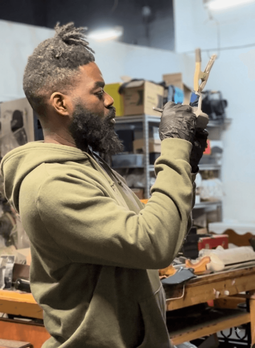 Man with a beard and dreadlocks wearing a hoodie and gloves, working with tools in a workshop.