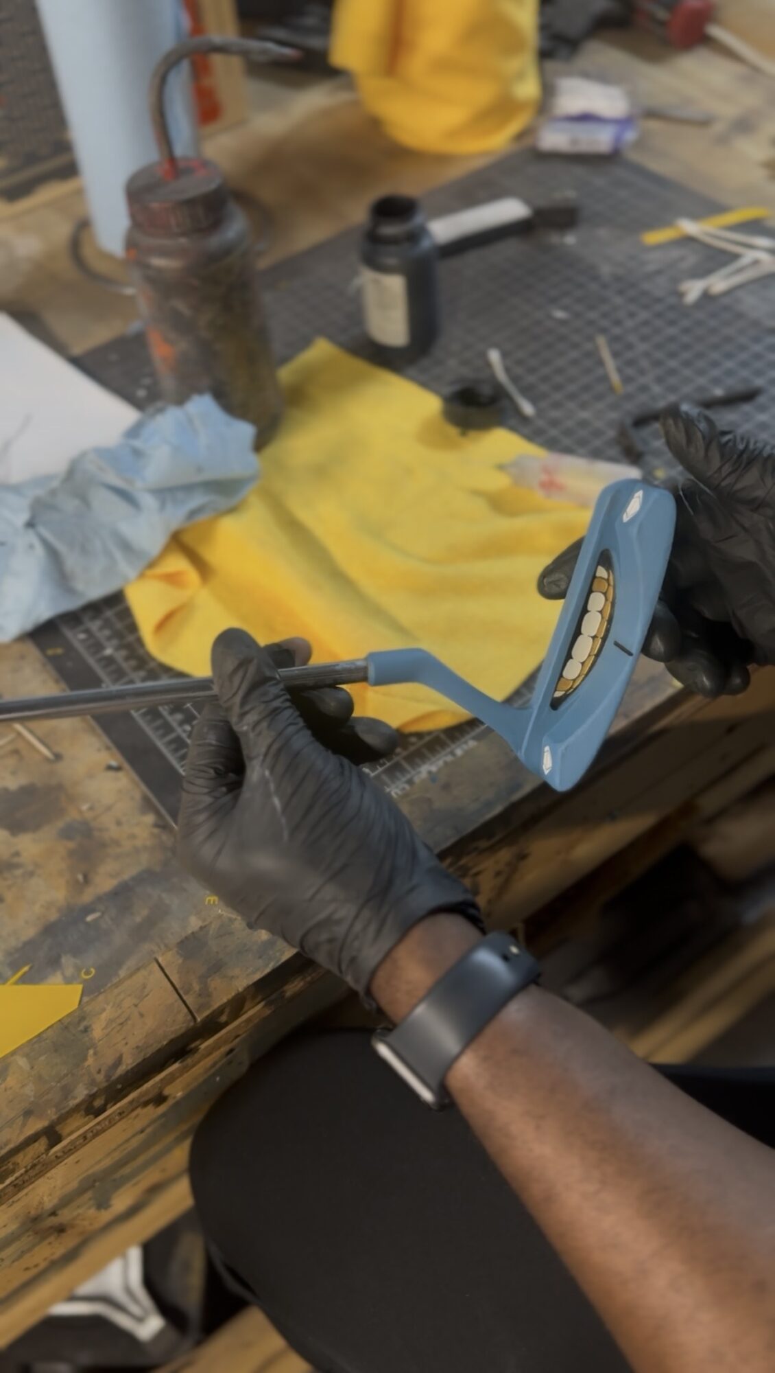 Person wearing black gloves holding a blue clamp on a workbench with tools and yellow material.