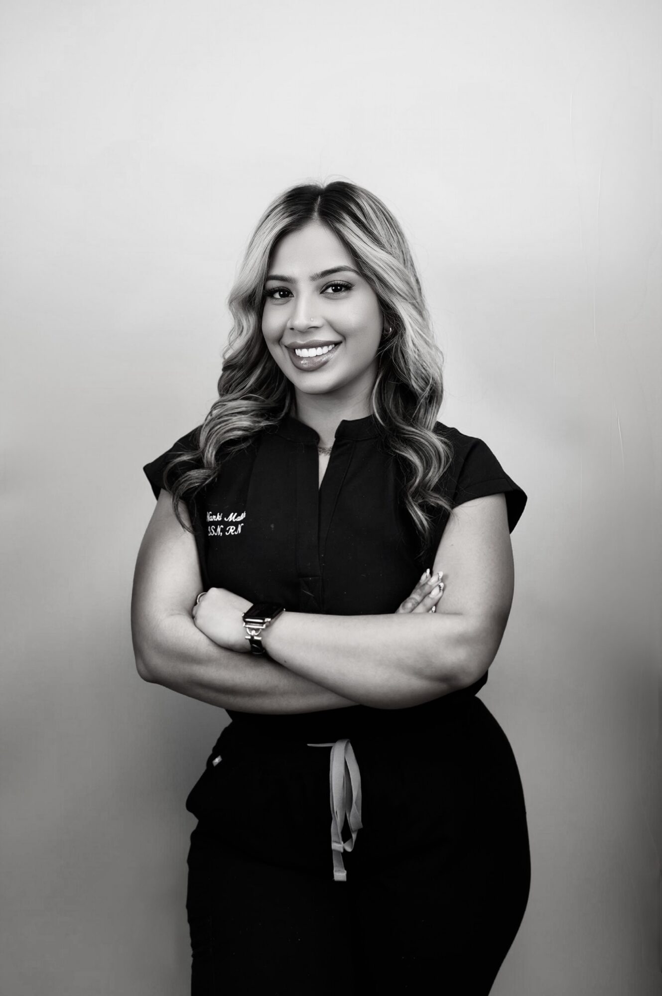 Young woman with wavy hair smiling, arms crossed, wearing a black shirt and pants, standing against a plain background.