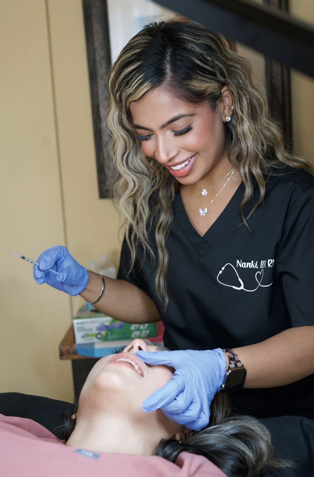 A healthcare professional with wavy hair and blue gloves administers a facial injection to a patient lying down.