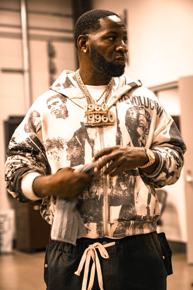 Man with a beard and jewelry wearing a patterned jacket and black pants, standing indoors.