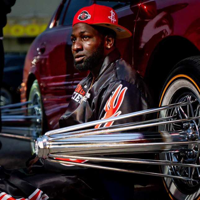 Man sitting next to a red car, wearing a red cap and black jacket, with metal rods in front. 20 words.