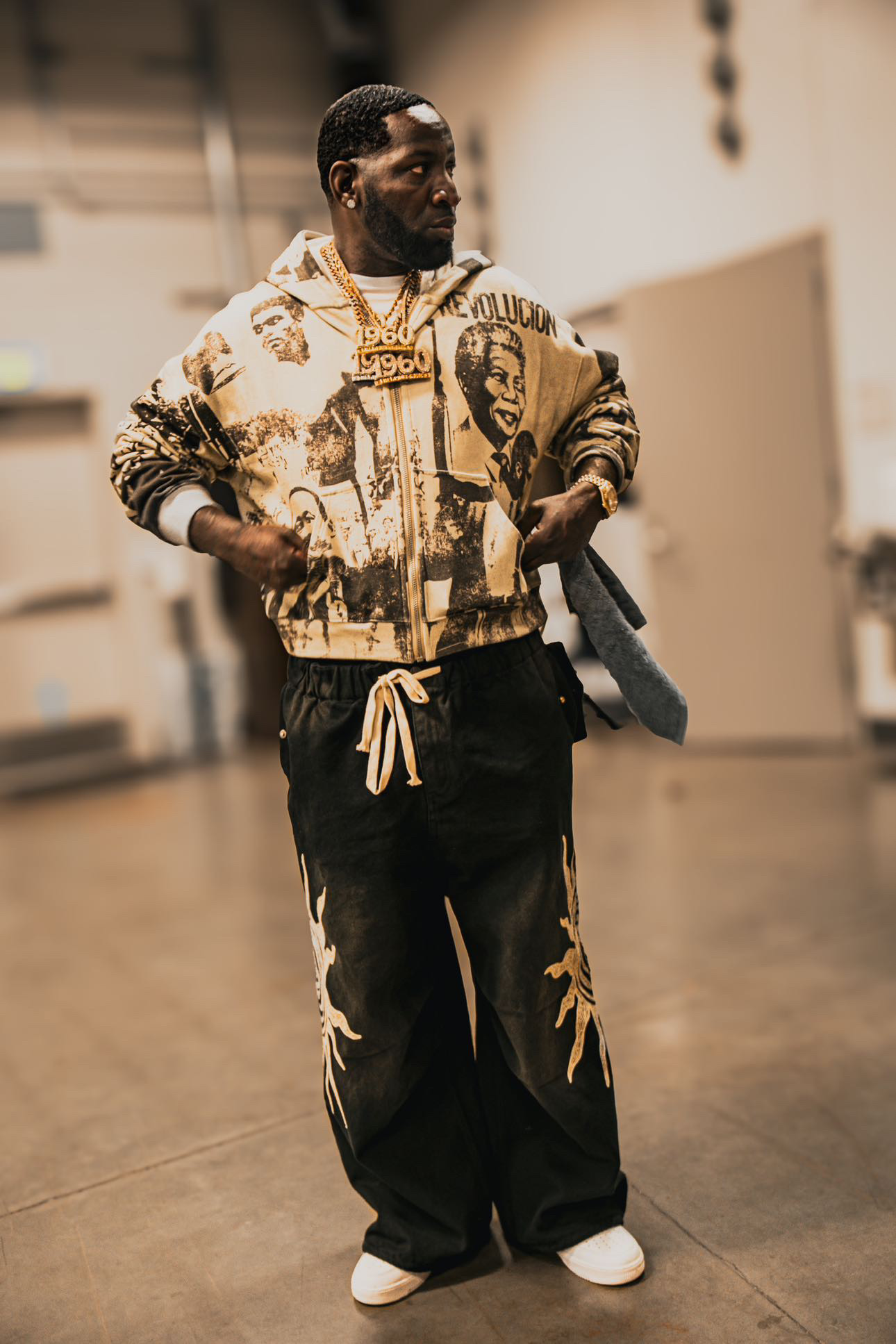 Man walking indoors wearing a patterned jacket and black pants, holding a towel, with a blurred background.