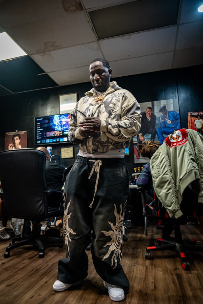Man standing in a gaming room with multiple monitors and chairs, wearing a beige jacket and black pants with floral designs.