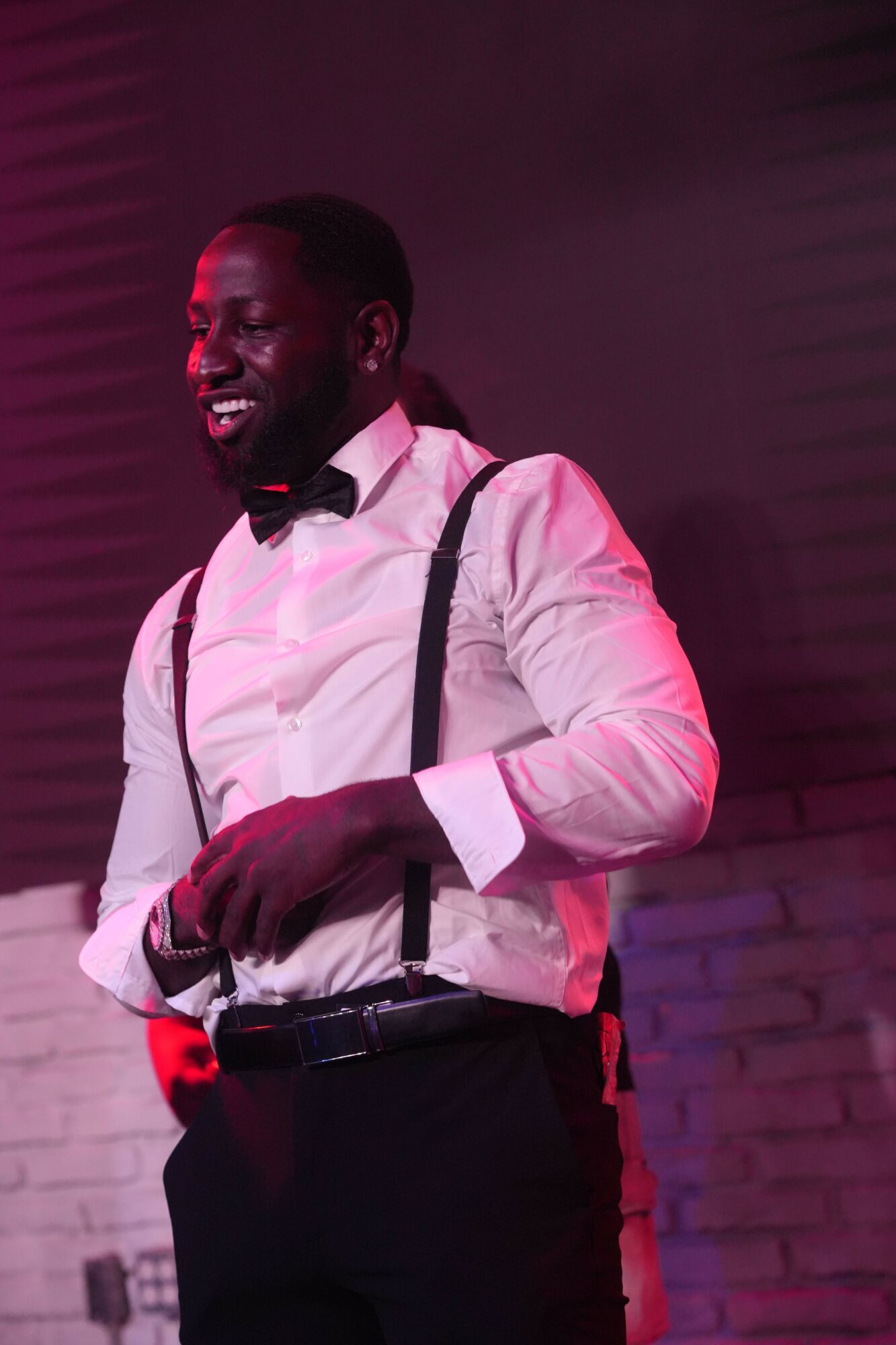 Man in white shirt and black pants smiling, standing with arms crossed, in a dimly lit room with pink lighting.