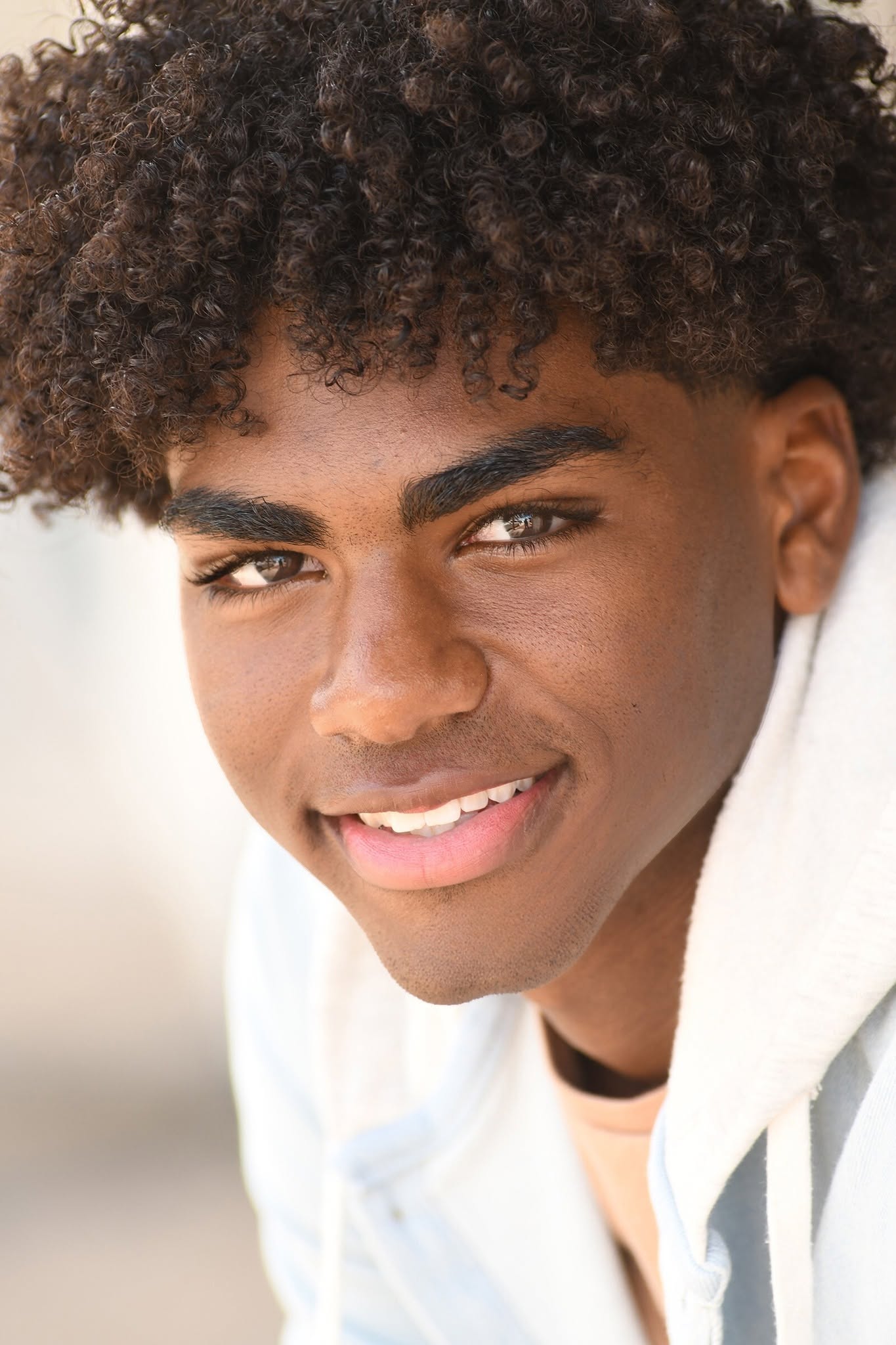 Close-up of a smiling young person with curly hair and dark eyes, wearing a white shirt.