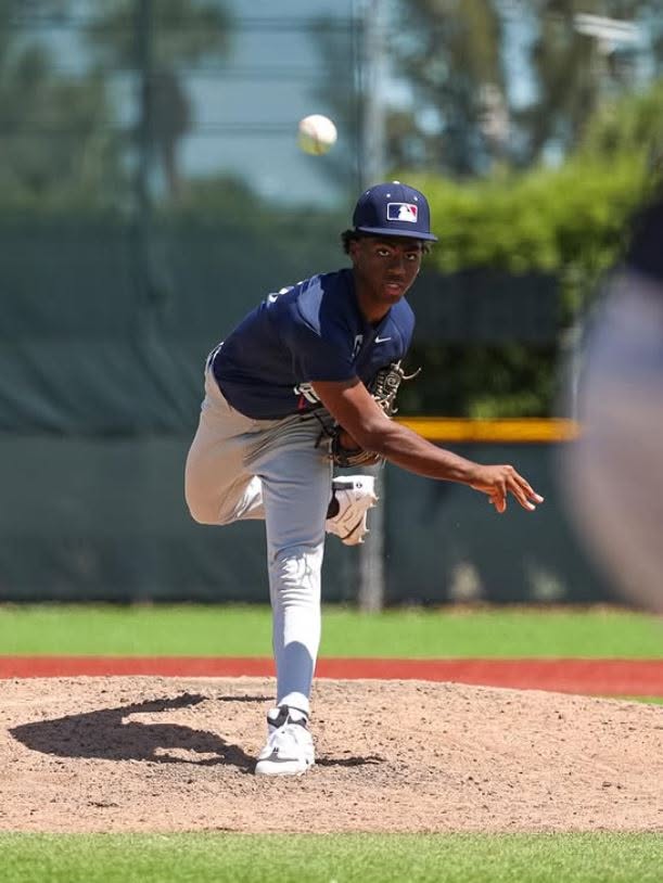 Baseball player in a navy uniform pitches on the field, with a baseball in mid-air, outdoors on a sunny day.