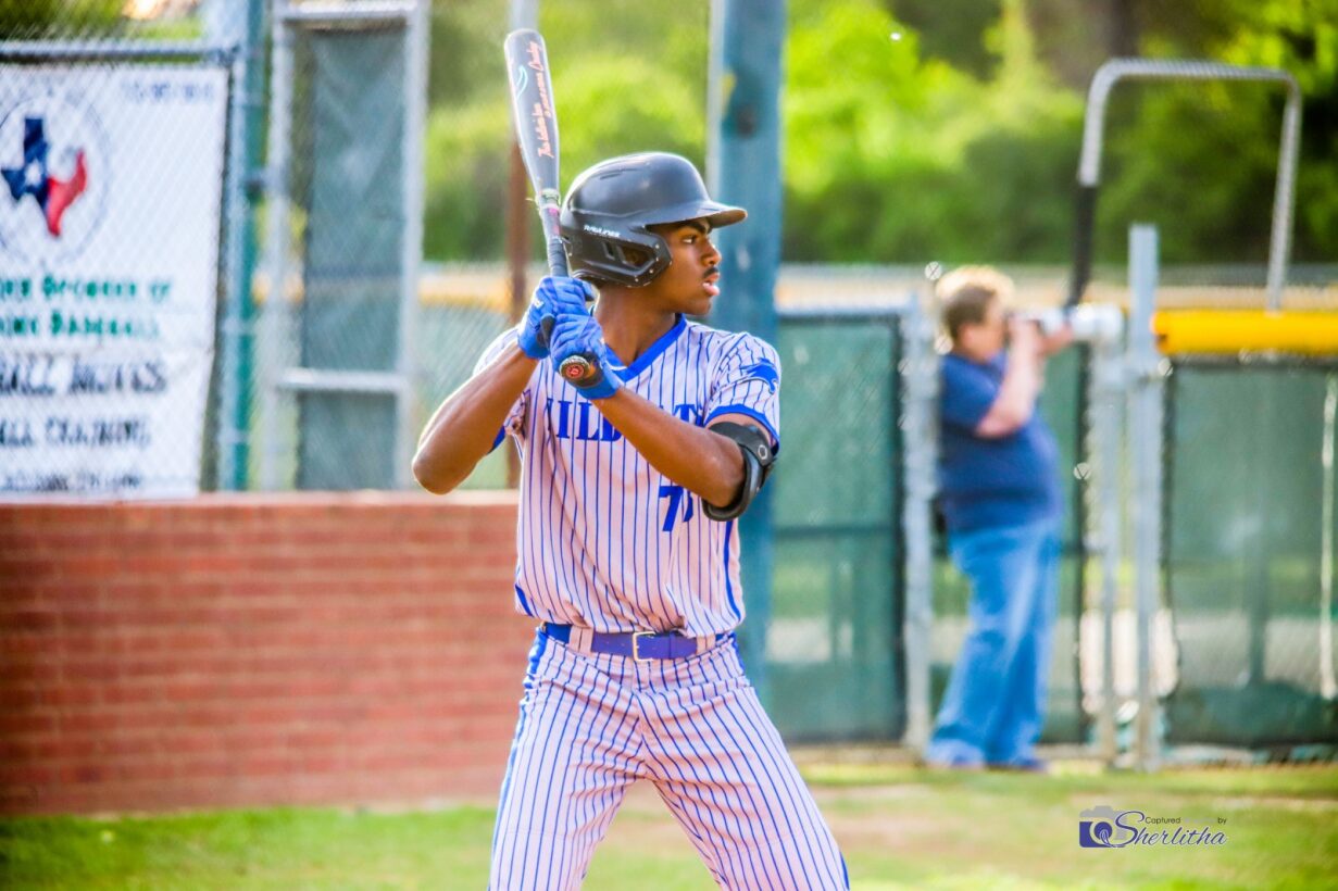 Young baseball player in striped uniform holding a bat, ready to swing, with a person in the background near a fence.