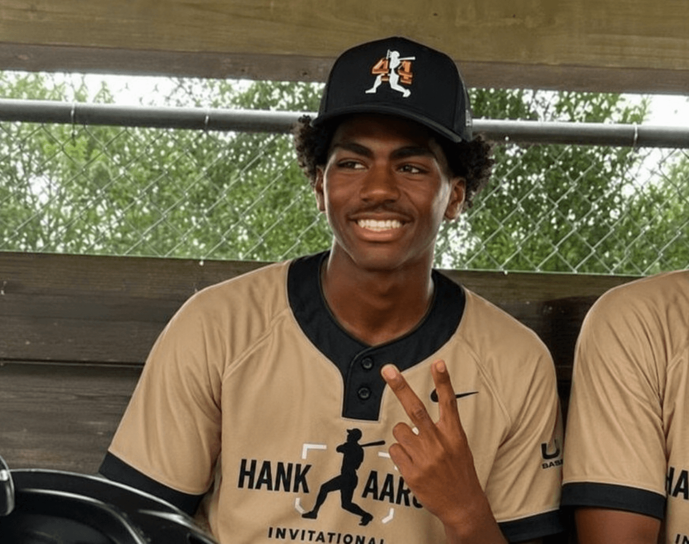 Young man smiling, wearing a black cap and beige sports jersey, making a peace sign with his fingers.
