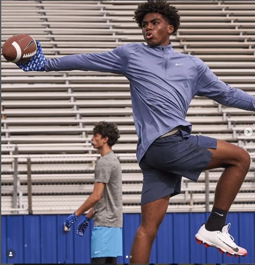 Young man in athletic clothing throws a football on a stadium field, with another person in the background. Empty bleachers are visible.
