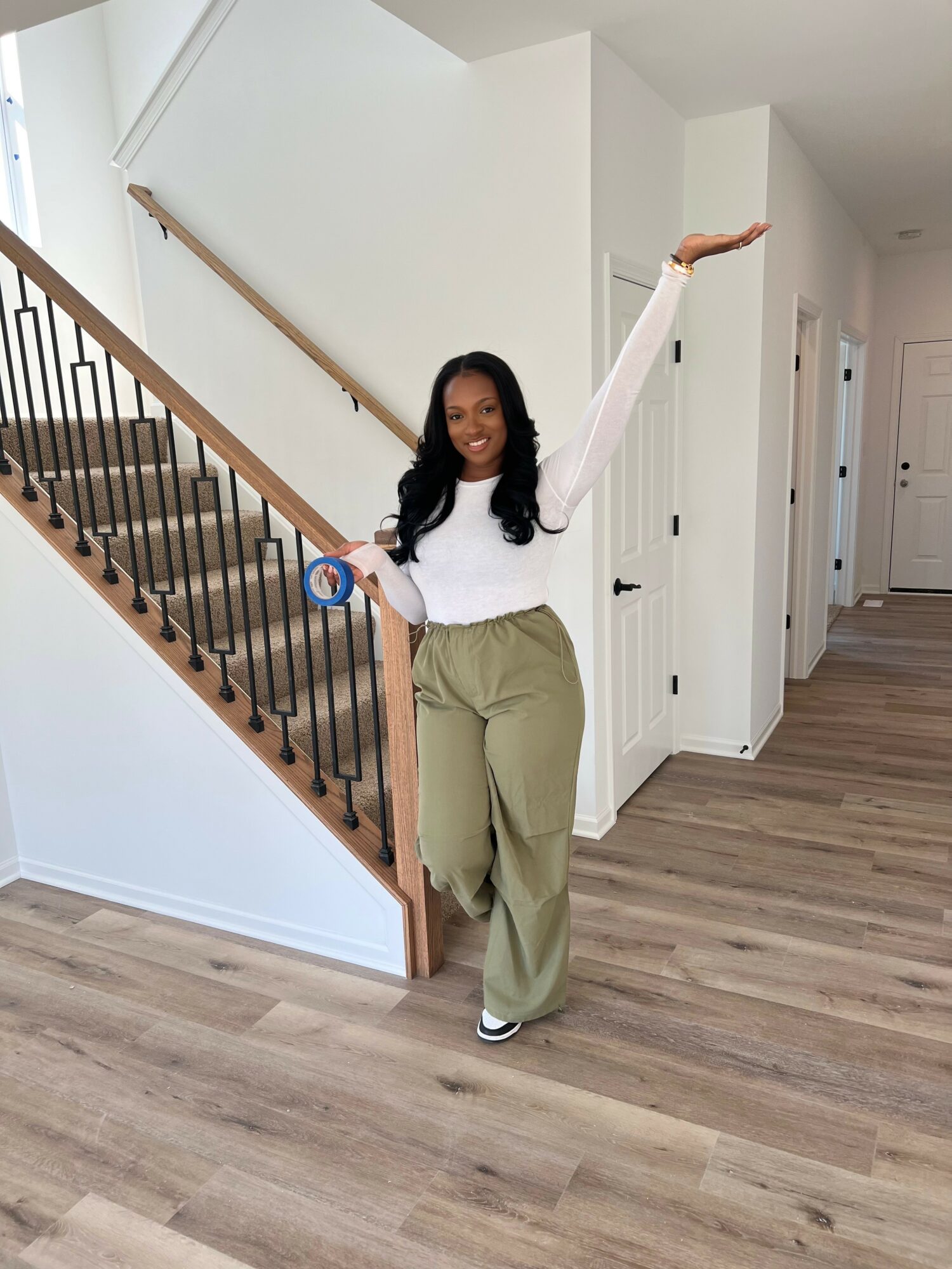 Woman standing indoors, smiling, with one arm raised, holding a small object, in a modern home with wooden floors and staircase.