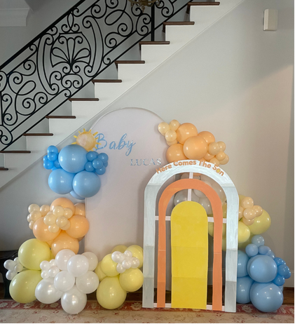 Balloon decoration with pastel colors and a rainbow arch, celebrating a baby's arrival, under staircase, with a sign reading 'Baby'.