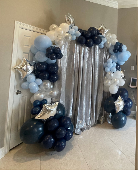 Balloon arch with silver curtains backdrop, featuring blue, navy, white, and silver balloons, some star-shaped, arranged around a doorway.