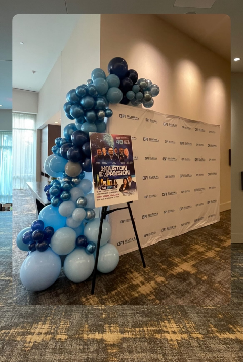Balloon arch with blue, black, and white balloons next to a promotional poster on an easel, in front of a backdrop with event branding.