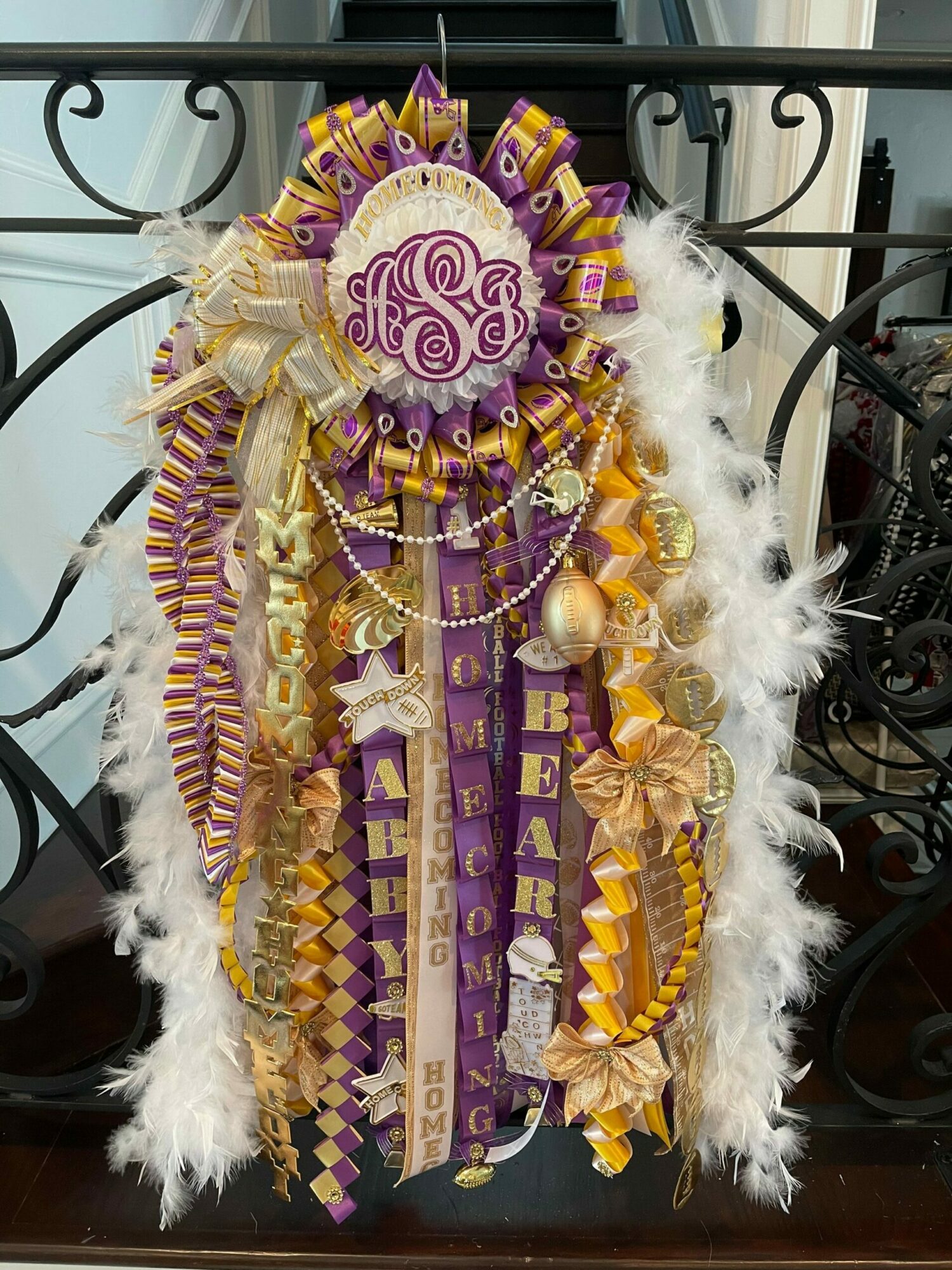 Decorative Mardi Gras float with purple, gold, and white ribbons, feathers, and a central emblem, displayed on a black metal stand.