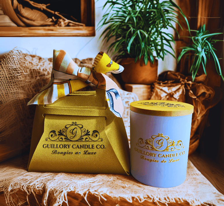 A yellow gift box with a ribbon and a dog-shaped ornament, and a white candle container with gold lettering, on a textured cloth, with plants in the background.