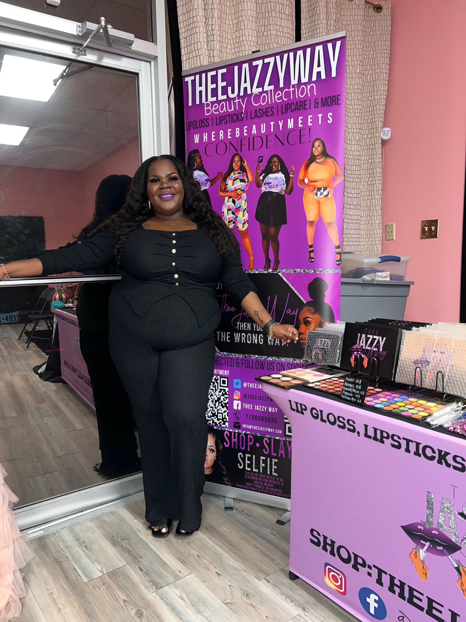 Woman standing next to a display table with beauty products, pink wall, and promotional poster in background.