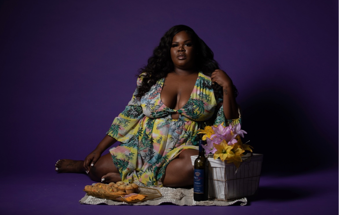 Woman sitting on a purple background with flowers, a bottle, and food, wearing a colorful dress, looking at the camera.