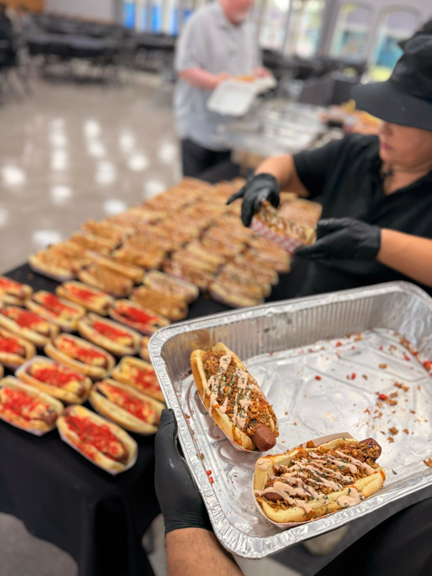 Person holding a tray with two hot dogs topped with sauce and toppings, with a table of hot dogs in the background.