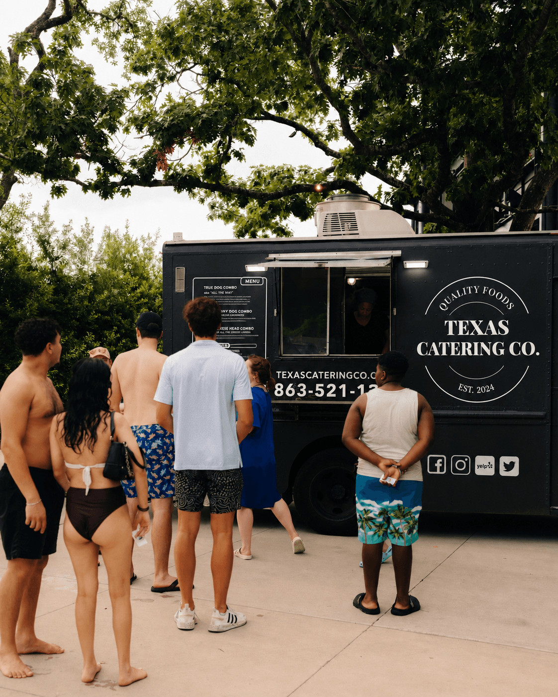 Group of people standing outside near a catering truck with trees in the background.