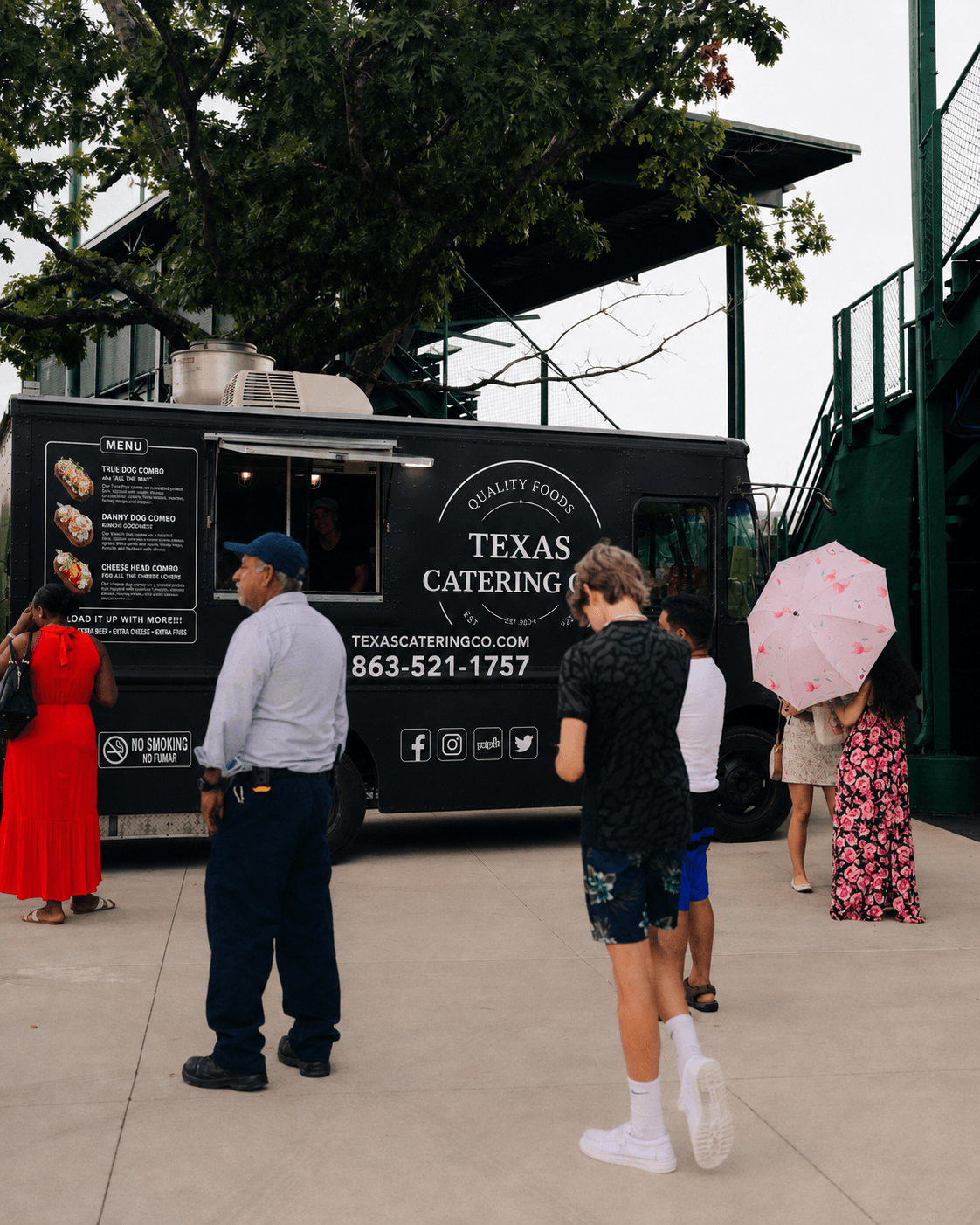 People standing in line outside a Texas catering food truck, some holding umbrellas, with trees and a building in the background.