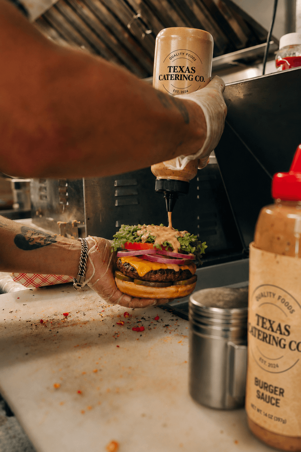 Person's arm pouring sauce onto a burger with lettuce, tomato, and meat, on a counter with condiments and kitchen tools.