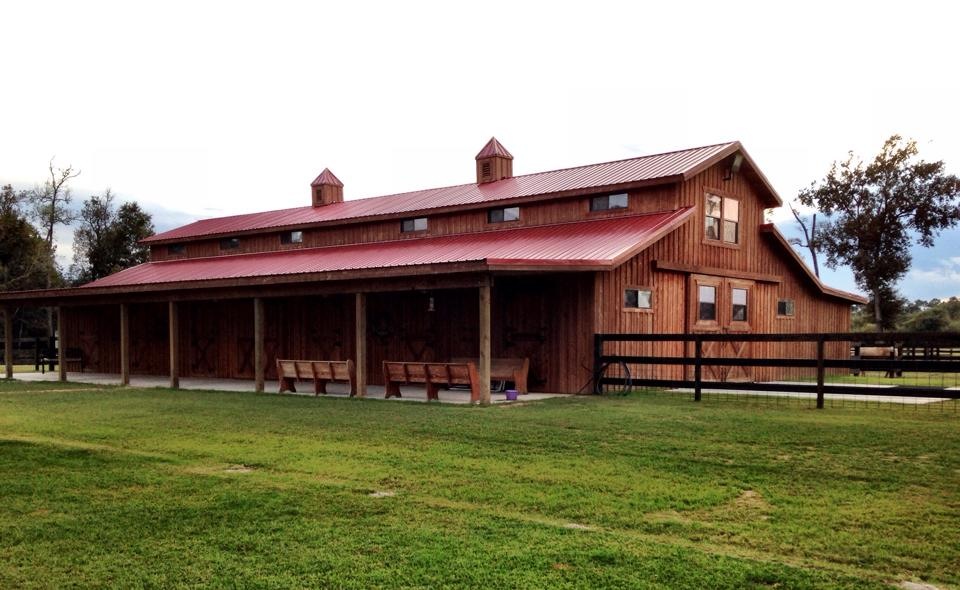 A large wooden barn with a red roof, surrounded by green grass and trees, under a cloudy sky.