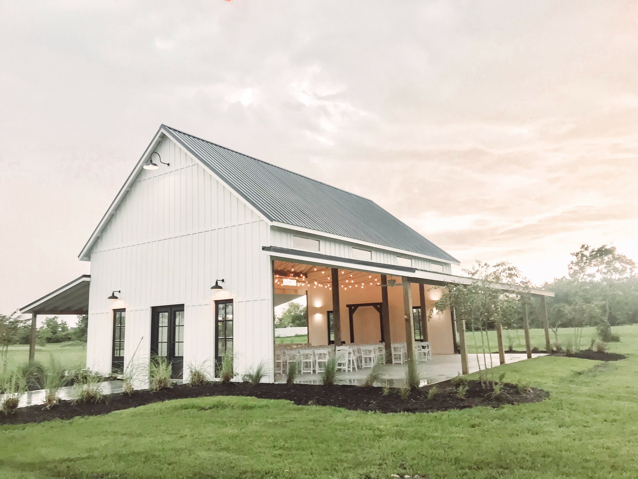 White barn-style building with large windows and a metal roof, surrounded by green grass and trees, under a cloudy sky.