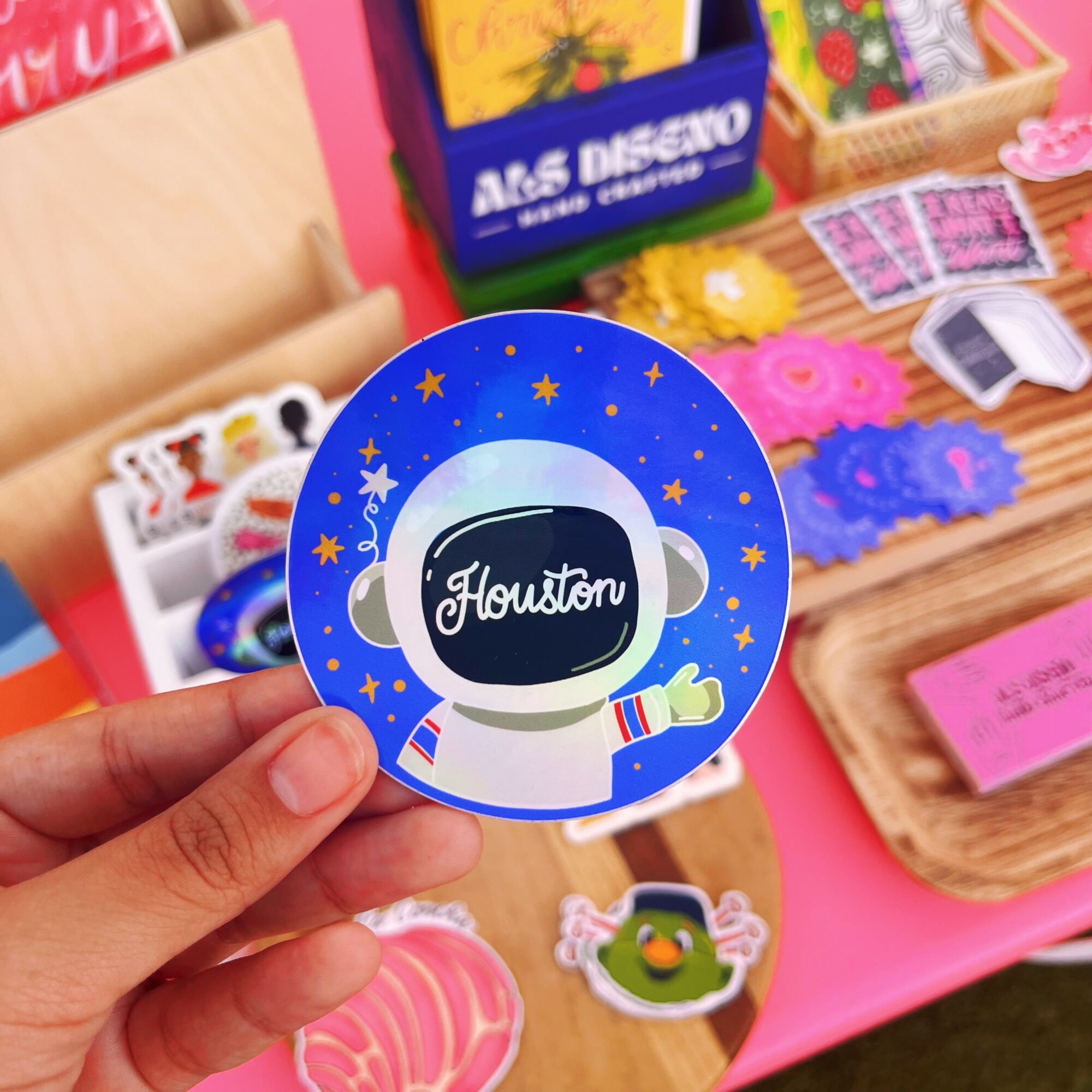 Hand holding a round sticker with a space-themed astronaut design and the word 'Houston' in the center.