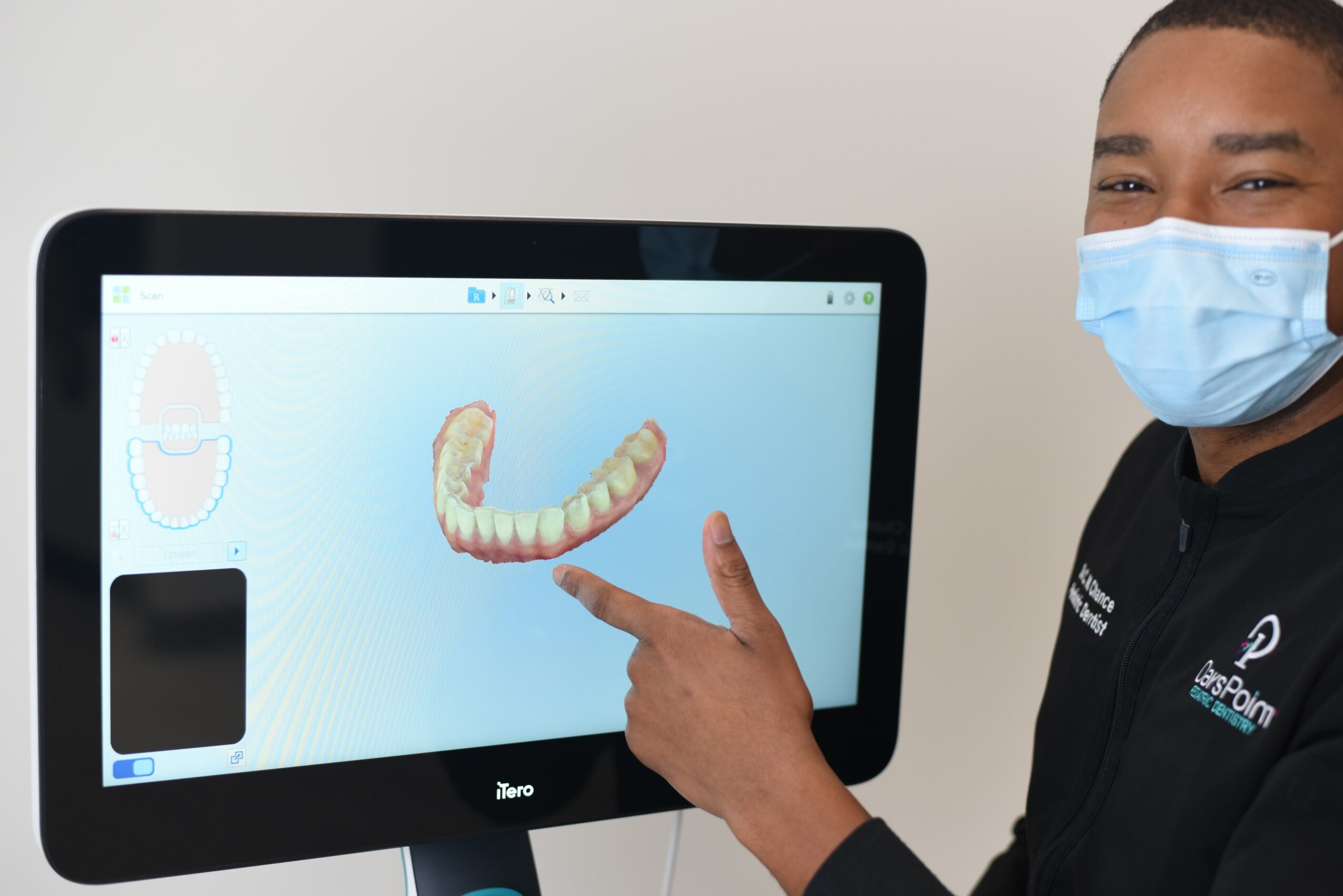 Person wearing a face mask pointing at a digital dental model on a computer screen, showing a set of teeth.