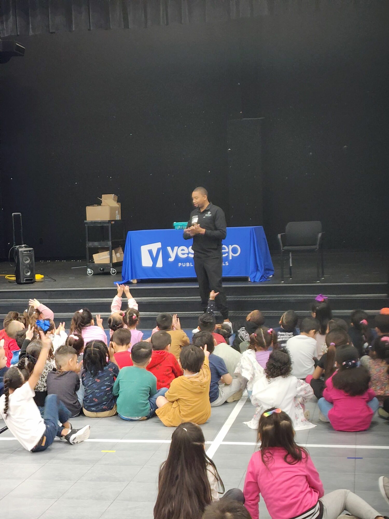 A person stands on stage in front of a large audience of children seated on the floor, with a black background and a blue sign.