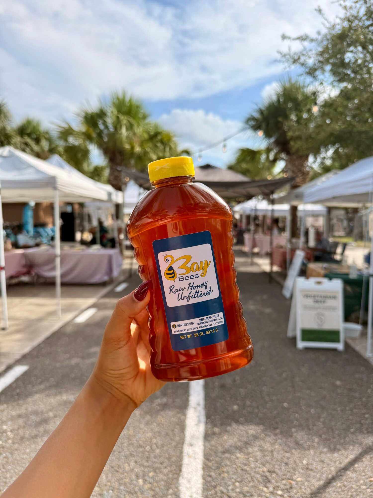 Hand holding a bottle of honey at an outdoor market with tents and trees in the background.