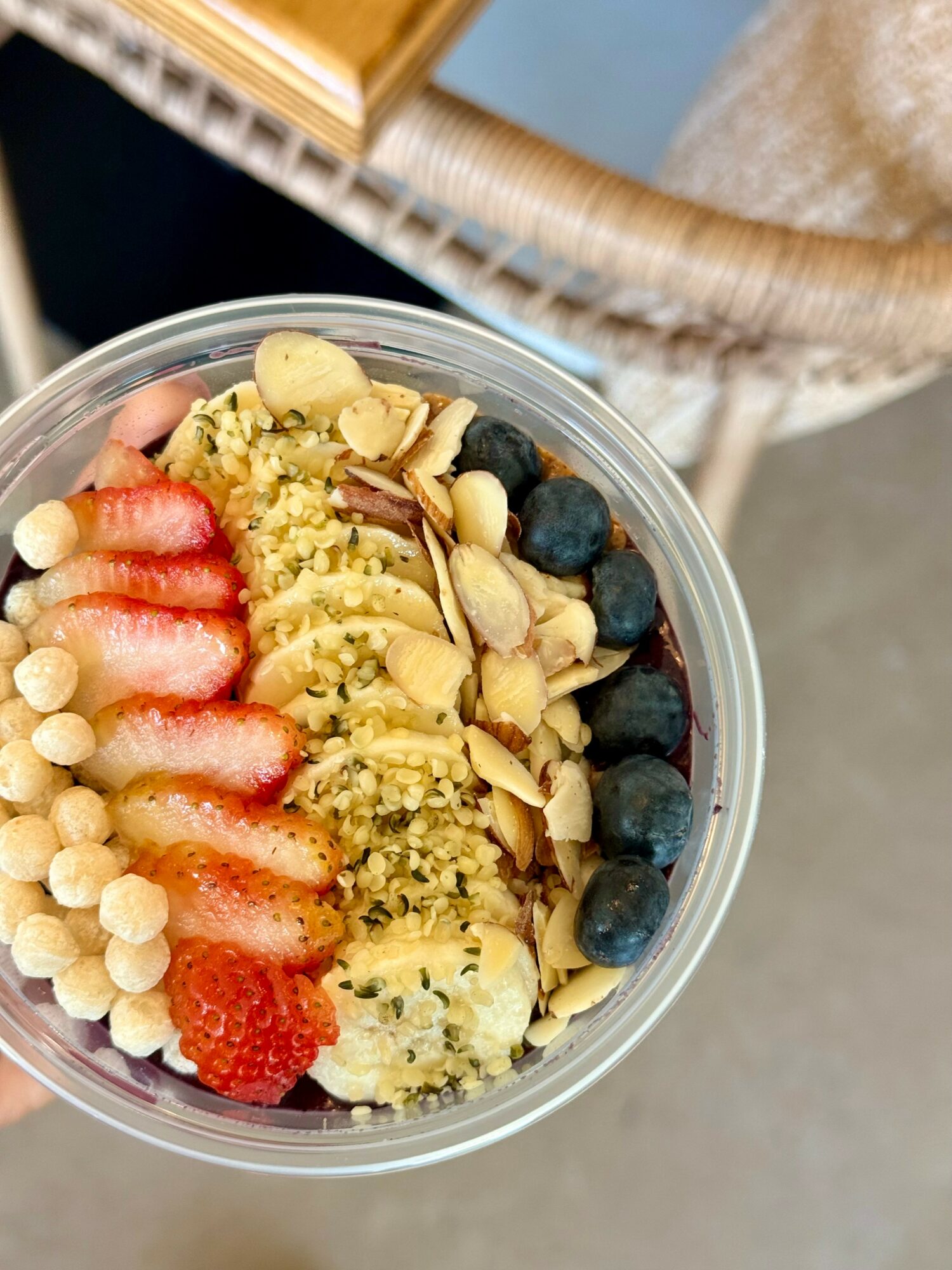 A bowl of yogurt topped with strawberries, blueberries, sliced almonds, and granola, held above a chair and table.
