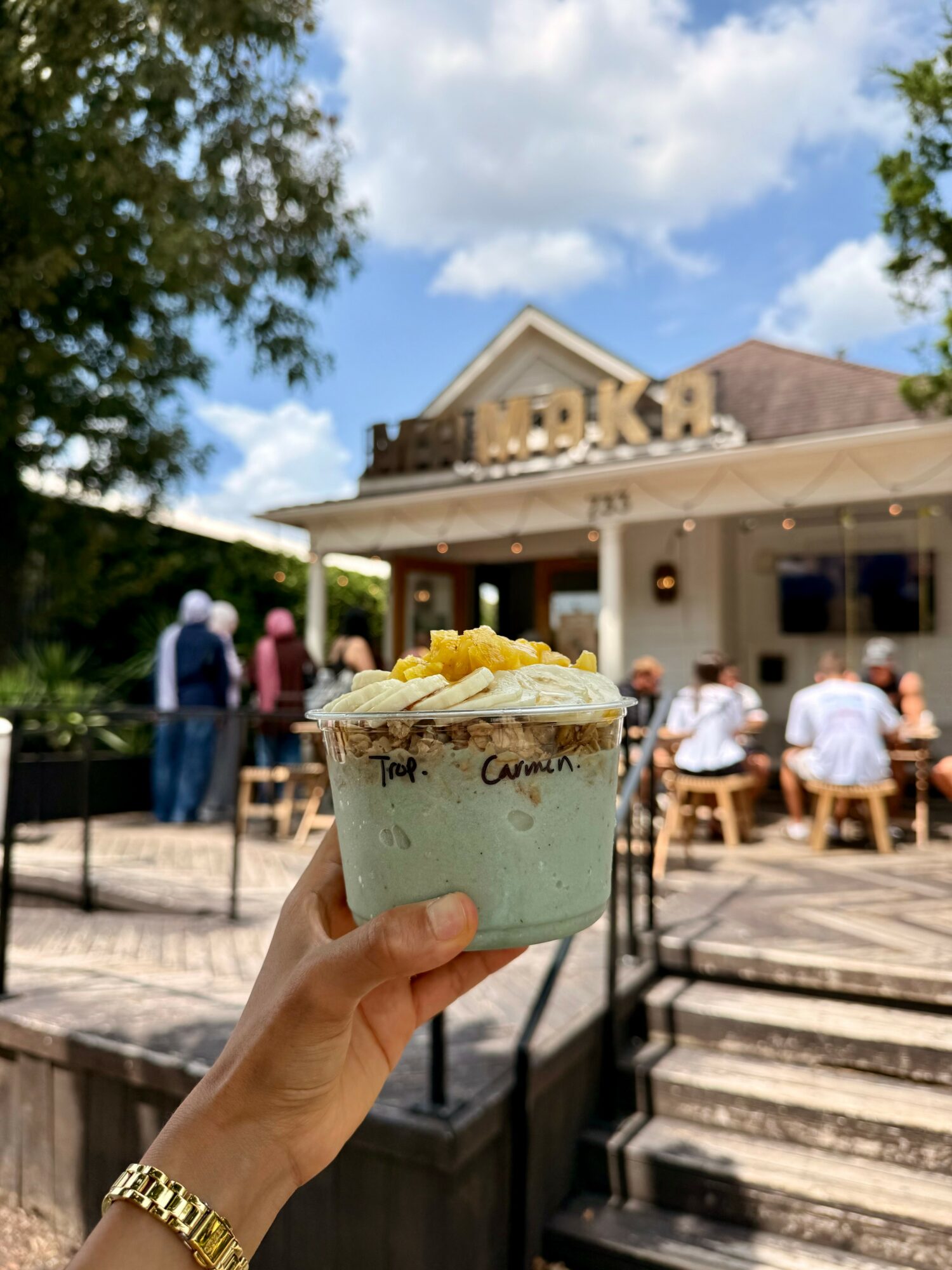 Hand holding a bowl of ice cream outdoors with a building and people in the background.