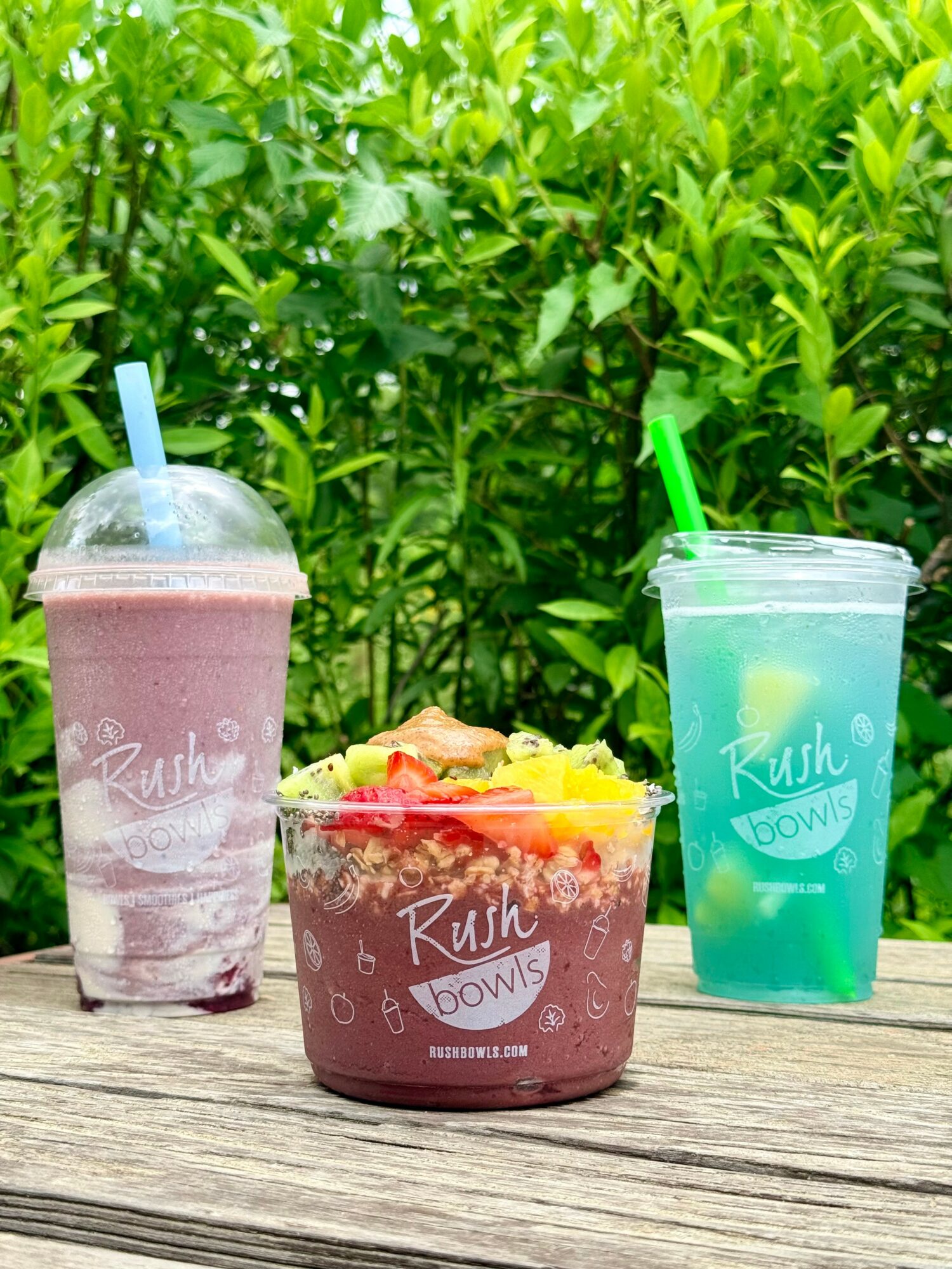 Two colorful smoothies and a bowl of fruit on a wooden surface with green foliage background.