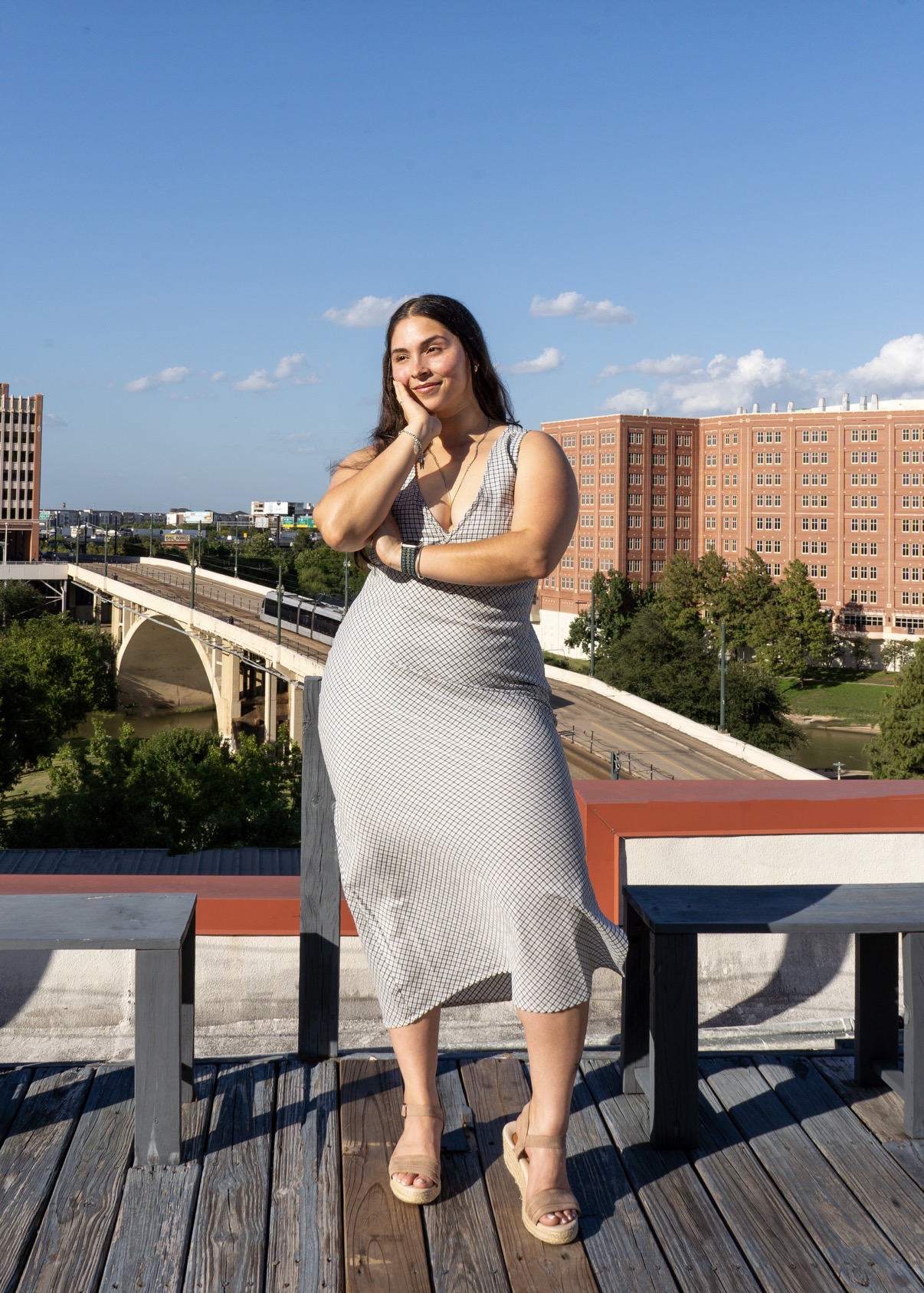 Woman standing outdoors on a wooden deck, smiling, with a cityscape and bridge in the background.