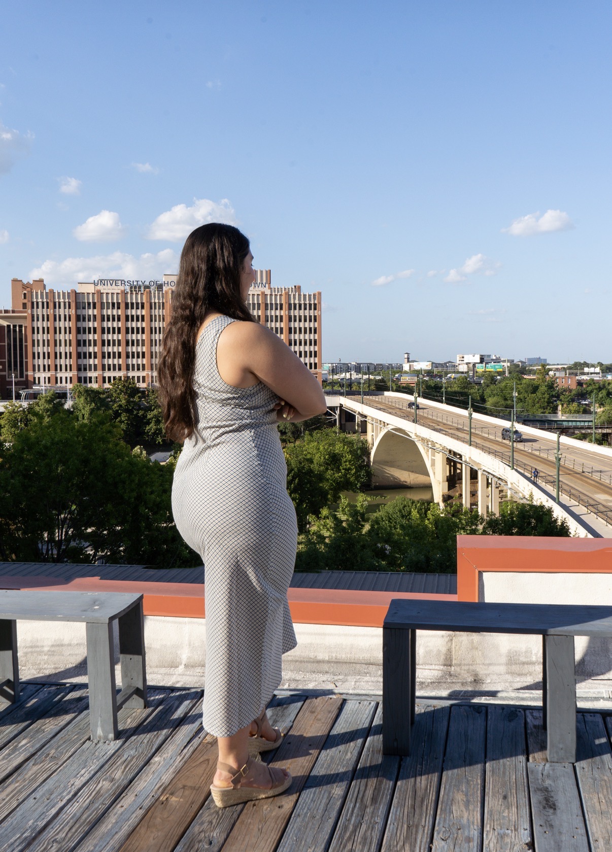 Woman with long hair in a sleeveless dress stands on a rooftop deck overlooking a cityscape with a bridge and buildings under a blue sky.
