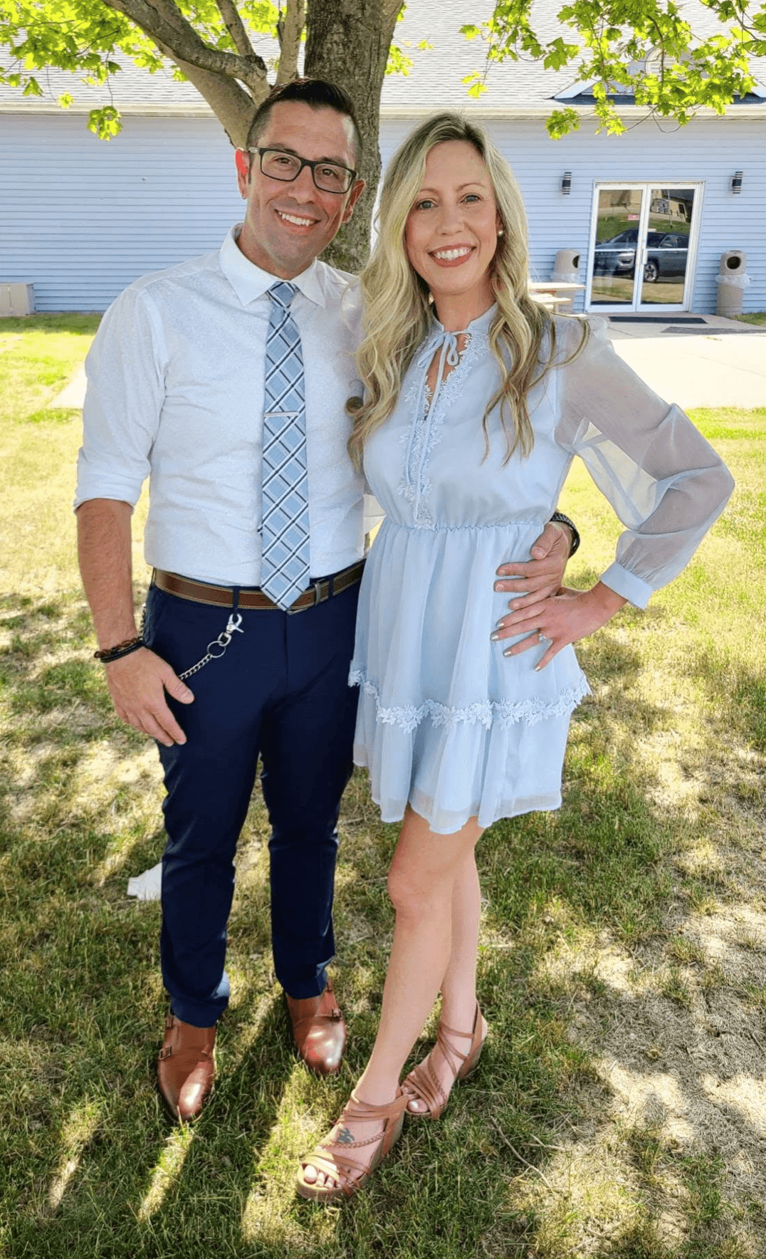 A man and woman stand outdoors on grass, smiling, with a house and tree in the background.