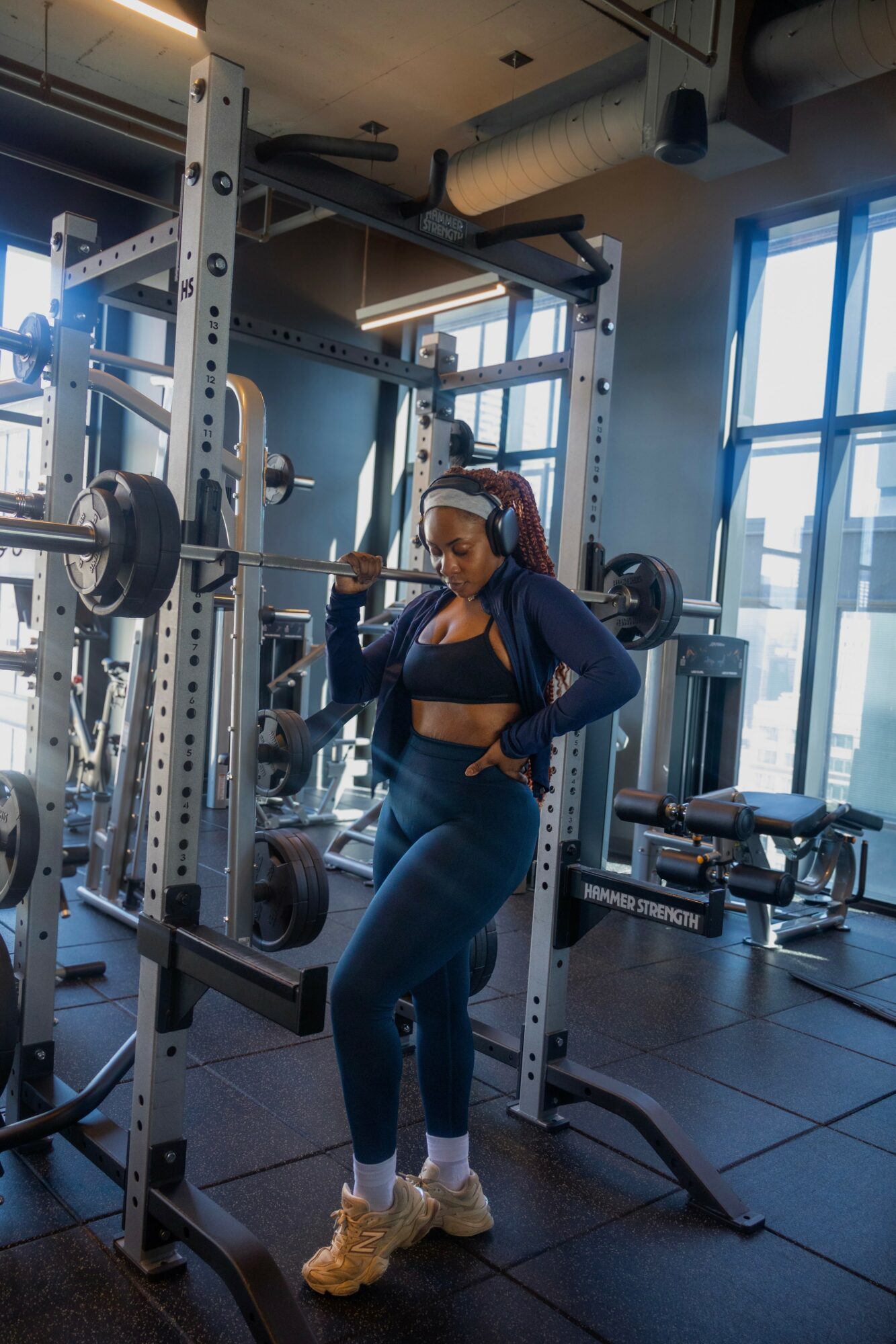 Woman in workout clothes standing in gym, holding barbell on shoulders, with weightlifting equipment around her.