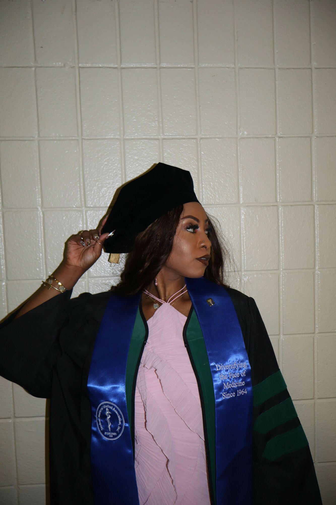 Person in graduation gown and cap making a peace sign, standing against a tiled wall.